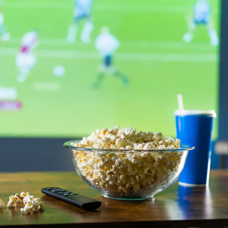 Popcorn and soda sitting on a coffee table in front of a 4K television with sport in the background